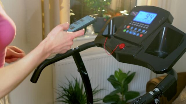 Running on treadmill while using smartphone unrecognizable woman during indoor home cardio workout. Holding mobile device and interacting with screen while jogging on electric fitness machine near