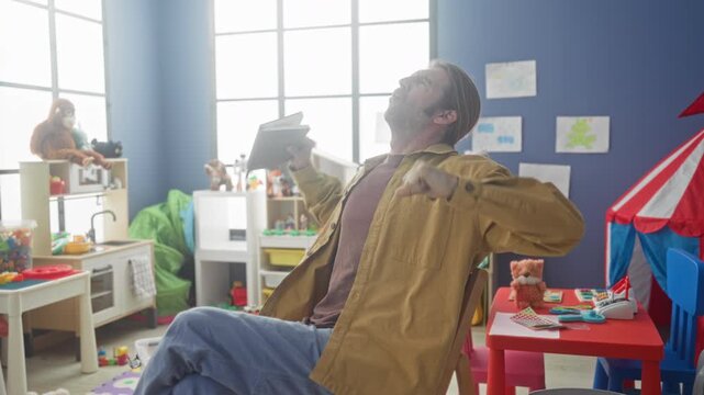 Man seated stretching and yawning while holding a book at a children's red table in a preschool playroom inside a building; tiredness.