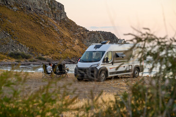 Fototapeta premium Family relax in folding chairs next to their modern campervan on a rugged beach in Limnos, Greece, enjoying a sunset view of the Aegean Sea during an off-grid motorhome road trip.