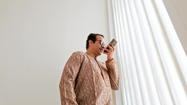 Man in an embroidered kurta speaks into a smartphone near tall window blinds in a bright, minimal interior.