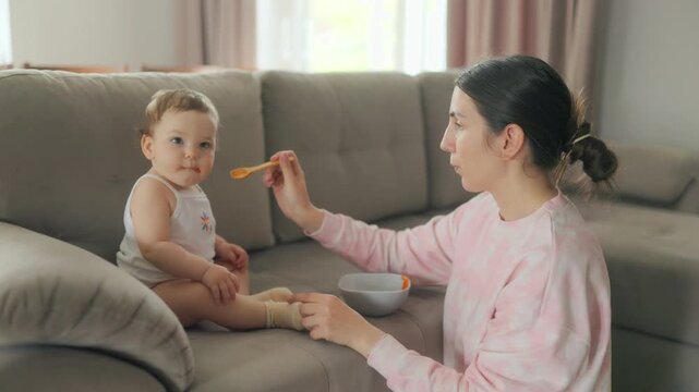 Side view of mother feeding infant baby with healthy puree on couch