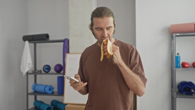 Man holding peeled banana and checking smartphone in a yoga studio, thumb scrolling while mid bite; quiet focus.