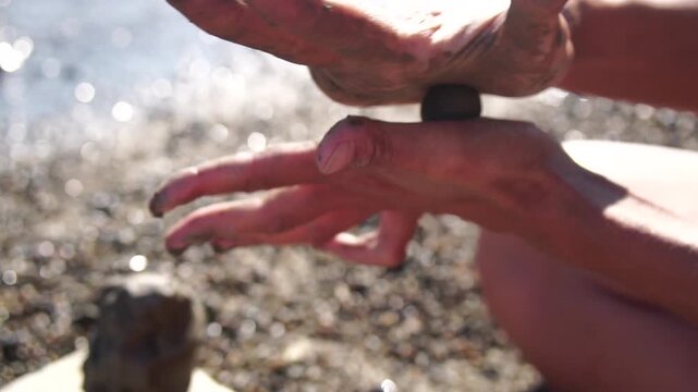 Hands mud ball: woman's dirty hands shaping a small ball of natural clay on a sunny pebble beach by the water in slow motion