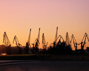 Obraz premium Industrial harbor cranes silhouetted against a glowing sunset sky over calm waterfront, creating a dramatic port landscape with maritime infrastructure, evening light and urban industrial atmosphere.