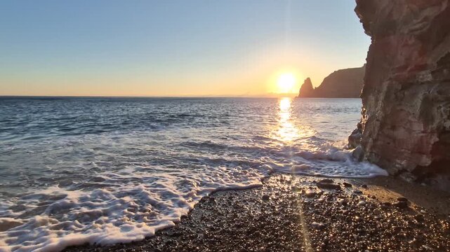 Sunset beach waves rolling over pebbles at the foot of dramatic coastal cliffs under a golden sky