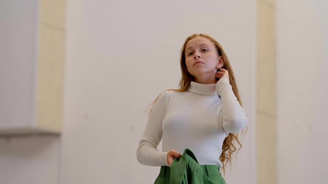 Teenage girl in white turtleneck putting on green blazer, getting dressed in classroom before school theatre rehearsal or performance.