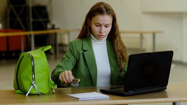 Young woman in glasses and green suit working on laptop like junior office worker or intern, sitting alone at table in coworking style room, concept freelance work and youth career start.