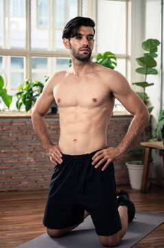 Fit athletic man kneeling on exercise mat after home workout. Concept of body fitness, strength training, healthy lifestyle, sport exercise, muscle development, and personal wellness.