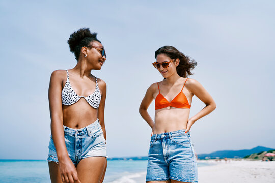 Two diverse young women in bikini tops and denim shorts stand on a sunny beach. They smile and share a moment, enjoying their summer vacation by the ocean.