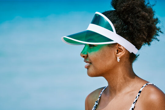 A young Black woman with curly hair wears a green transparent visor and a patterned swimsuit. She looks to the left, enjoying a bright, sunny day outdoors against a blue sky.