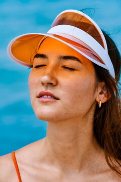 A young woman with closed eyes wears an orange visor, enjoying a sunny day outdoors. Her face is tinted orange by the visor. She wears a swimsuit top.