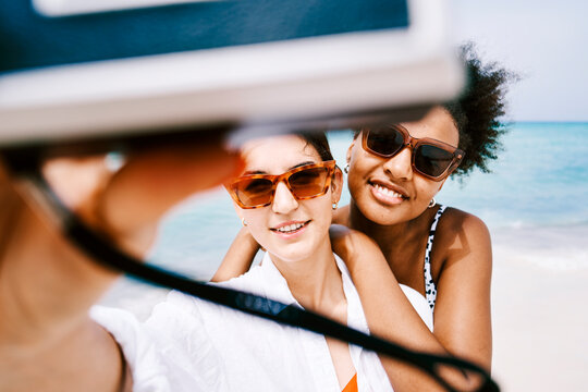 Two smiling young women, friends on vacation, wear sunglasses and take a selfie on a beautiful sandy beach. The clear blue ocean provides a stunning backdrop for their happy moment.