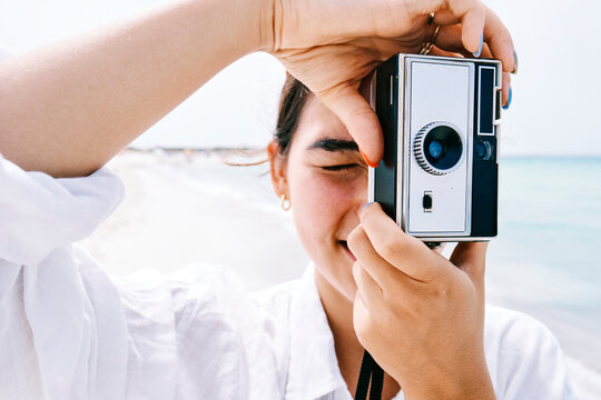 A young person in a white shirt holds a retro camera, framing a shot on a bright, sunny beach. Their eyes are closed, concentrating on capturing the perfect vacation memory.