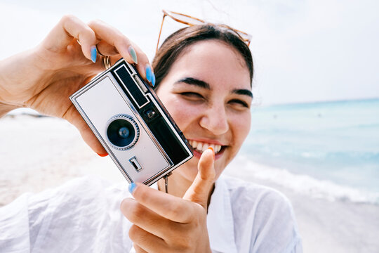 A happy young woman holds a vintage camera, aiming it at the viewer on a sunny beach. She smiles broadly, enjoying her vacation by the ocean.
