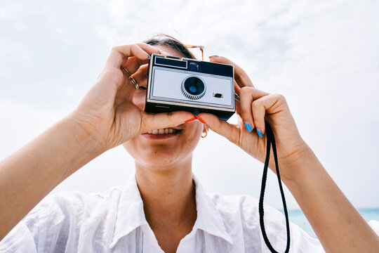 A smiling young woman holds a vintage camera up to her face, looking through the viewfinder. She is enjoying a bright day outdoors, capturing memories with her classic film camera.