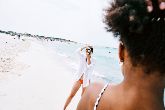A young woman photographs her friend on a beautiful white sand beach with clear blue water. They are enjoying a sunny day by the ocean. Other beachgoers are in the background.