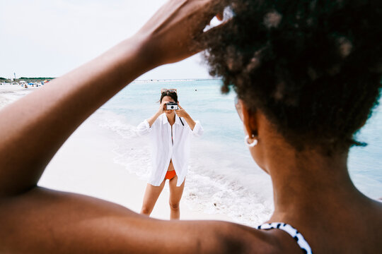 A woman on a beach photographs her friend, captured from a first-person perspective. The photographer stands by the ocean, while the subject's back and arm are visible in the foreground.