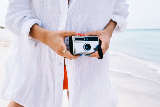 A woman wearing a white linen shirt holds a vintage film camera on a sunny beach. Her hands with gold rings and bright nail polish are ready to capture photos of the ocean.