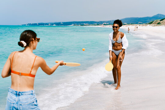 Two young women play paddleball on a sunny beach. One woman in a bikini smiles, holding a paddle. The other hits the ball near the turquoise ocean.