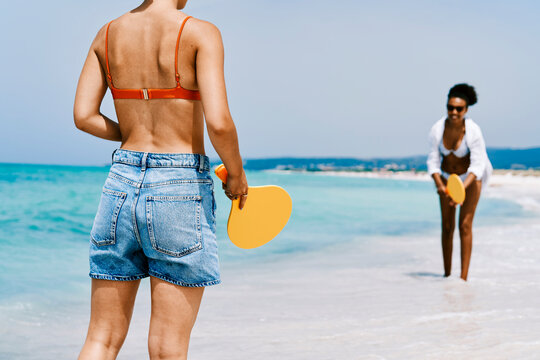 Two women play paddleball on a sunny beach with turquoise water. One woman stands with her back to the camera, holding a yellow paddle. Another woman stands in the distance, ready to play.
