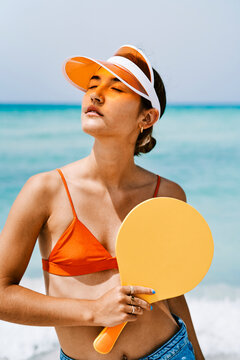 A young woman in an orange bikini top and visor stands on a sunny beach, holding a yellow paddle. She closes her eyes, enjoying the warm sun and ocean breeze.