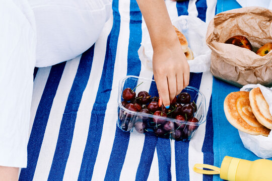 A person's hand reaches for fresh cherries from a plastic container on a striped picnic blanket. Other snacks like pastries and fruit are nearby for an outdoor meal.