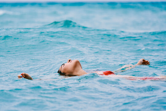 A woman in a red swimsuit floats peacefully on her back in the calm blue ocean water. Her eyes are closed, enjoying a moment of relaxation and tranquility.