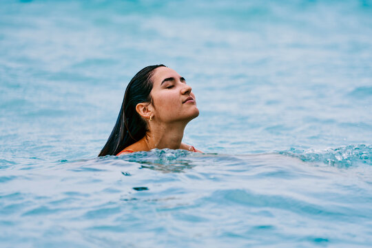 A young woman with wet dark hair enjoys a refreshing swim in clear blue water. Her eyes are closed as she relaxes, feeling the cool water.