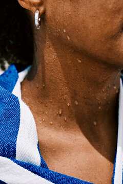 A close-up of water droplets on the neck of a person wearing a blue and white striped towel and a silver earring. The subject is wet from water on a sunny day.