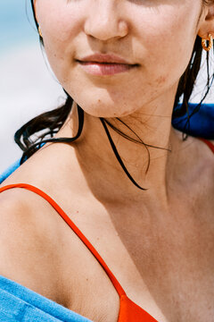 A young woman with damp hair from swimming stands in the sunlight. She wears a vibrant red bikini strap and a blue towel while enjoying a calm summer day at the coast.