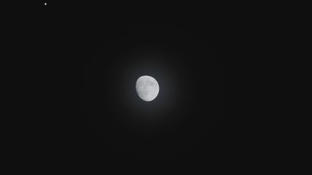 Airplane flies across dark night sky, its silhouette crossing in front of a bright gibbous moon. Aircrafts navigation lights are visible as it passes by this celestial event