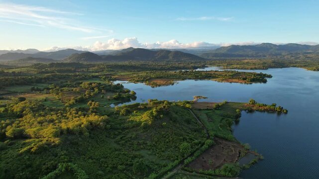 Aerial drone footage of the edge of Batu Bulan water reservoir lake, at early sunrise, in Sumbawa regency, Sumbawa island, Indonesia, with wild green nature and mountains behind