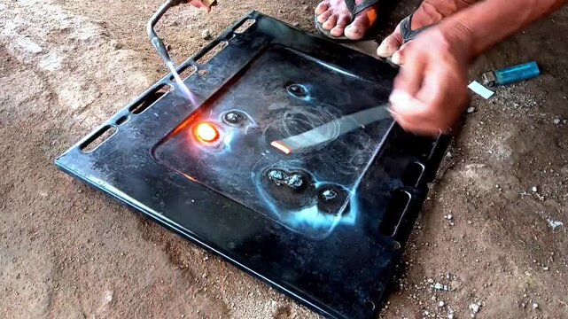 Hands apply a lit gas torch and a silver metal rod to a black rectangular panel, creating bright orange sparks and light on dirt ground near a small, blue plastic handheld lighter.