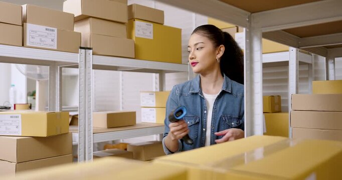 Asian woman entrepreneur standing in warehouse aisle while checking shipping label on parcel box, reviewing inventory shelf position, managing online shopping parcel box workflow inside storage area