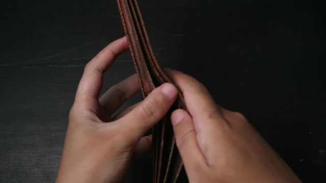 Hands holding and opening an empty brown leather wallet against a dark wooden background showing financial crisis.