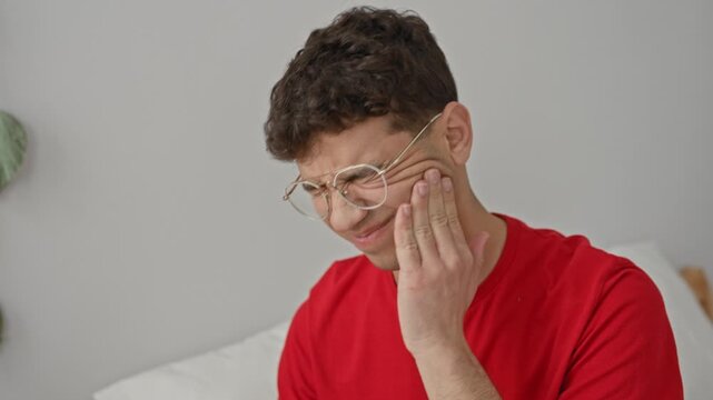 Man touching cheek, hand on cheek for toothache while wincing on a bed in a building interior, wearing glasses; toothache pain.