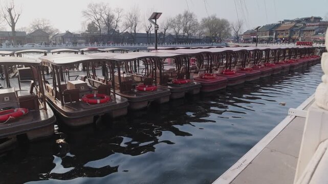 Traditional wooden boats with red cushions moored in a row along Houhai Lake waterfront in Beijing. Static wide angle shot captures winter scenery with bare trees and historic hutong buildings.