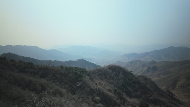 Mutianyu's Jundu Mountains stretch across the frame with layered ridges fading into winter haze. Slow lateral pan reveals the Yan Range topography with sparse vegetation on hillsides.
