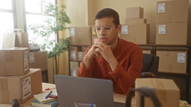 Man in a small business building clasps hands under chin over laptop amidst stacked boxes and a scanner on desk, wearing glasses and looking pensive; anxiety.