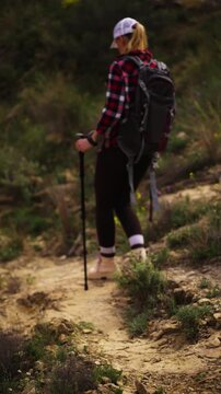 Woman with backpack and trekking poles hiking on narrow mountain trail. Determined trekker walking downhill on dirt path surrounded by green vegetation during sunny day