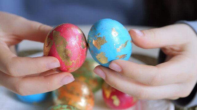 Hands hold colorful Easter eggs with gold foil patterns. Background shows more decorated eggs. Focus on vibrant red and blue eggs. Soft lighting highlights glossy textures