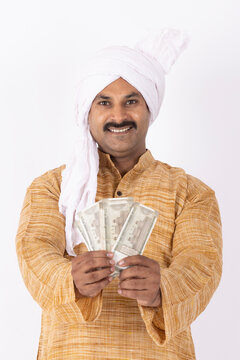 Portrait of smiling young farmer showing indian rupees currency wearing kurta pajama and white turban gesturing while standing against white background
