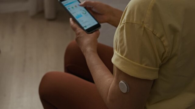 Over shoulder shot of middle aged woman using glucose monitoring app while wearing CGM sensor on upper arm during routine diabetes care at home, tilt down camera movement