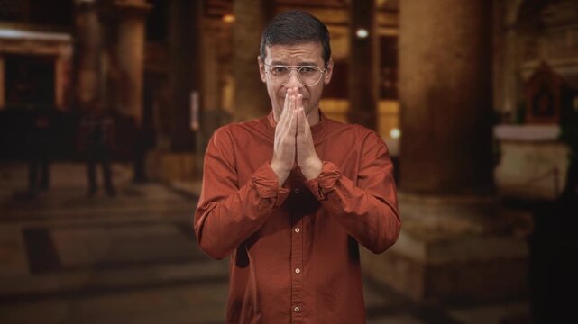 Young hispanic man praying with hands pressed together, wearing glasses and a rust shirt inside an ornate building interior with stone columns; penitence reflection.