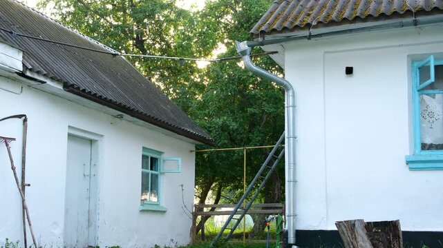 Simple whitewashed village house with light blue windows and modest roof stands beside a small shed. Quiet summer countryside scene showing authentic rural lifestyle and tradition.
