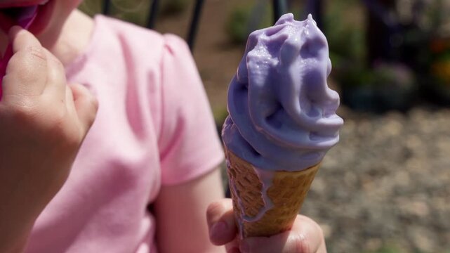 Close up of a little girl in a pink shirt eating delicious purple lavender ice cream in a waffle cone with a pink spoon. Child enjoying a cold lilac summer treat in a sunny outdoor park garden
