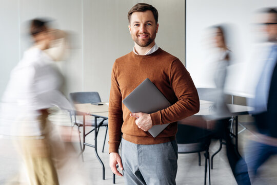 Confident businessman standing with laptop, smiling at camera as blurred colleagues walk past in a modern office environment