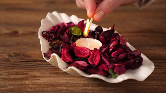 Close-up of a hand lighting a small tea light candle placed inside a white decorative shell filled with vibrant pink and red dried flower petals. The scene is set on a rustic wooden table, evoking a s