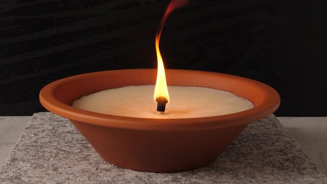 Burning memorial candle in a terracotta bowl on a stone base. Bright flame flickering against a dark background. Symbol of remembrance, mourning, and commemoration during All Saints' Day. 4K video.
