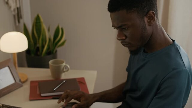 Tilt up shot of focused young adult Black man browsing websites on laptop sitting at desk in home office while wearing CGM patch on upper arm, daily diabetes care concept